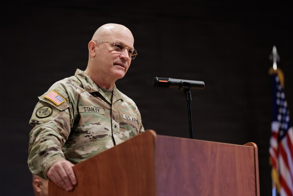 A U.S. Army Brigadier General named Stanley is speaking at a wooden podium with a microphone. He is wearing the Army Combat Uniform (ACU) in MultiCam pattern with a U.S. flag patch on the right shoulder and a single black star indicating his rank of Brigadier General. An American flag is visible in the background to the right.