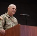 A U.S. Army Brigadier General named Stanley is speaking at a wooden podium with a microphone. He is wearing the Army Combat Uniform (ACU) in MultiCam pattern with a U.S. flag patch on the right shoulder and a single black star indicating his rank of Brigadier General. An American flag is visible in the background to the right.