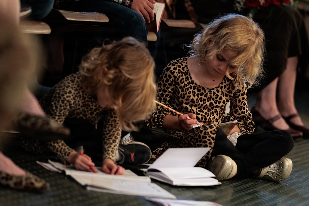 Two young children with blonde curly hair, both wearing matching leopard-print outfits, are sitting on the floor writing or drawing in notebooks. They appear to be in an auditorium or event space, with adults seated in chairs behind them. The older child is focused on writing with a pencil while the younger child uses a red crayon or marker.