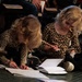 Two young children with blonde curly hair, both wearing matching leopard-print outfits, are sitting on the floor writing or drawing in notebooks. They appear to be in an auditorium or event space, with adults seated in chairs behind them. The older child is focused on writing with a pencil while the younger child uses a red crayon or marker.