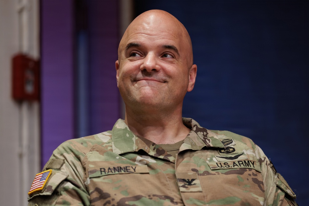 A U.S. Army soldier wearing an OCP (Operational Camouflage Pattern) uniform is smiling and looking upward to his right. The name tape on his uniform reads 'RANNEY' and the branch tape reads 'U.S. ARMY.' He appears to be in an indoor setting with a colorful background.