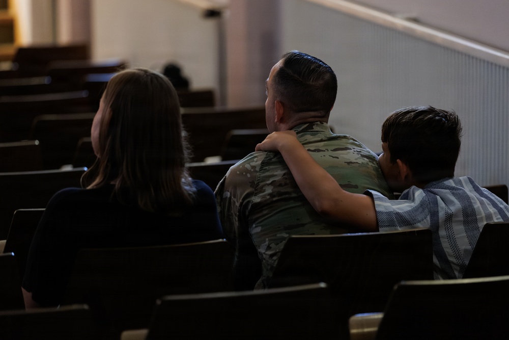 Three individuals are seated in wooden pews inside what appears to be a chapel or auditorium. The central figure is wearing military camouflage (OCP/MultiCam pattern) uniform, flanked by two others who appear to be embracing them. The scene suggests an emotional or supportive moment, photographed from behind.
