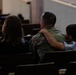 Three individuals are seated in wooden pews inside what appears to be a chapel or auditorium. The central figure is wearing military camouflage (OCP/MultiCam pattern) uniform, flanked by two others who appear to be embracing them. The scene suggests an emotional or supportive moment, photographed from behind.
