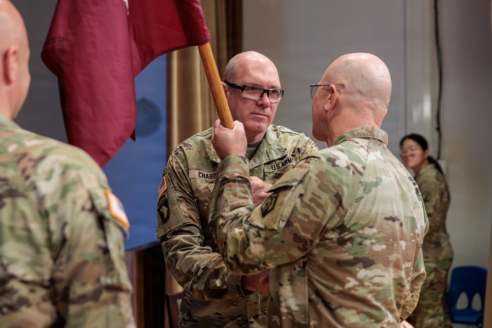 A U.S. Army military ceremony is taking place, likely a change of command or activation ceremony, where a guidon (unit flag) is being passed between soldiers. The central figure, identified by the name tape 'CHABO' (partially visible), is receiving or transferring the maroon/burgundy guidon. Multiple soldiers in OCP (Operational Camouflage Pattern) uniforms are present in what appears to be an indoor venue.
