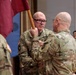 A U.S. Army military ceremony is taking place, likely a change of command or activation ceremony, where a guidon (unit flag) is being passed between soldiers. The central figure, identified by the name tape 'CHABO' (partially visible), is receiving or transferring the maroon/burgundy guidon. Multiple soldiers in OCP (Operational Camouflage Pattern) uniforms are present in what appears to be an indoor venue.