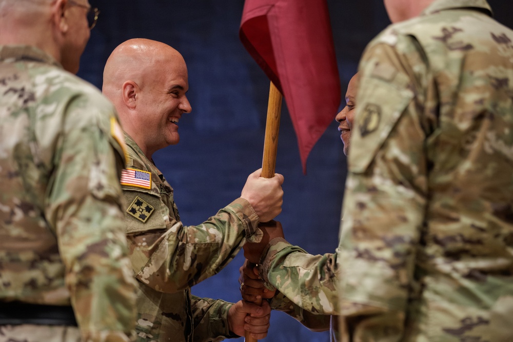 A military change of command ceremony is taking place, with two soldiers exchanging a guidon (unit flag) with a wooden staff. The central figure is a bald, smiling soldier wearing U.S. Army OCP (Operational Camouflage Pattern) uniform with a 4th Infantry Division patch and American flag patch. Additional uniformed soldiers are visible in the background and foreground, suggesting a formal military ceremony.