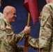 A military change of command ceremony is taking place, with two soldiers exchanging a guidon (unit flag) with a wooden staff. The central figure is a bald, smiling soldier wearing U.S. Army OCP (Operational Camouflage Pattern) uniform with a 4th Infantry Division patch and American flag patch. Additional uniformed soldiers are visible in the background and foreground, suggesting a formal military ceremony.
