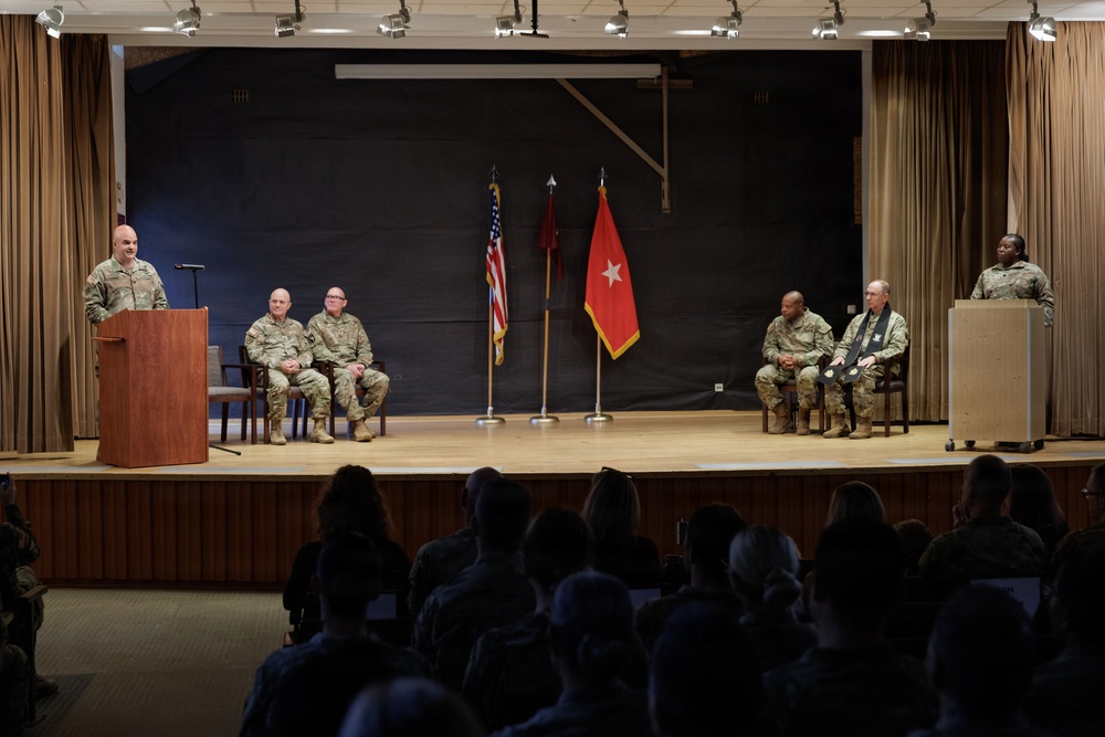 A formal U.S. Army military ceremony is taking place on a stage with six uniformed personnel. One soldier speaks at a podium on the left while another stands at a podium on the right, with four seated soldiers in between flanked by flags. A large audience of uniformed military personnel observes from the darkened foreground.