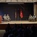 A formal U.S. Army military ceremony is taking place on a stage with six uniformed personnel. One soldier speaks at a podium on the left while another stands at a podium on the right, with four seated soldiers in between flanked by flags. A large audience of uniformed military personnel observes from the darkened foreground.