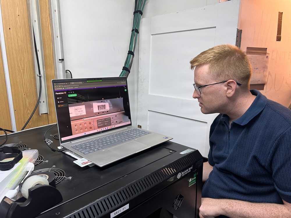A young man with red hair and glasses is seated at a workstation, intently viewing a laptop screen displaying what appears to be a web application called 'ParaLine' with a pink/salmon-colored industrial control interface. The laptop is resting on top of a Black Box rack-mount equipment unit labeled 'RR01.01'. The setting appears to be a technical or industrial workspace with cable management infrastructure visible in the background.