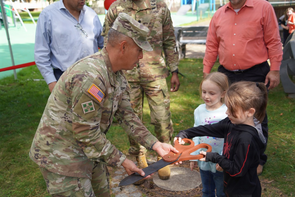 A U.S. Army soldier in OCP camouflage uniform is participating in a ribbon-cutting ceremony with two young children, using an oversized ceremonial scissors. The event appears to take place outdoors in a playground or park setting. Several other adults, including another soldier and two civilians, are observing in the background.
