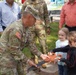 A U.S. Army soldier in OCP camouflage uniform is participating in a ribbon-cutting ceremony with two young children, using an oversized ceremonial scissors. The event appears to take place outdoors in a playground or park setting. Several other adults, including another soldier and two civilians, are observing in the background.