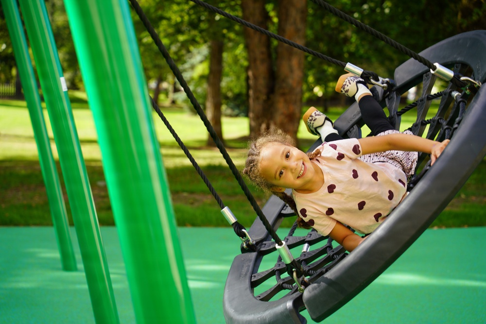 A young girl with curly hair is lying on a large nest-style swing at a modern playground, smiling at the camera. She is wearing a white top with heart patterns, dark leggings, and leopard-print shoes. The playground features bright green equipment and a rubberized safety surface, set against a backdrop of lush green trees.
