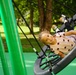 A young girl with curly hair is lying on a large nest-style swing at a modern playground, smiling at the camera. She is wearing a white top with heart patterns, dark leggings, and leopard-print shoes. The playground features bright green equipment and a rubberized safety surface, set against a backdrop of lush green trees.