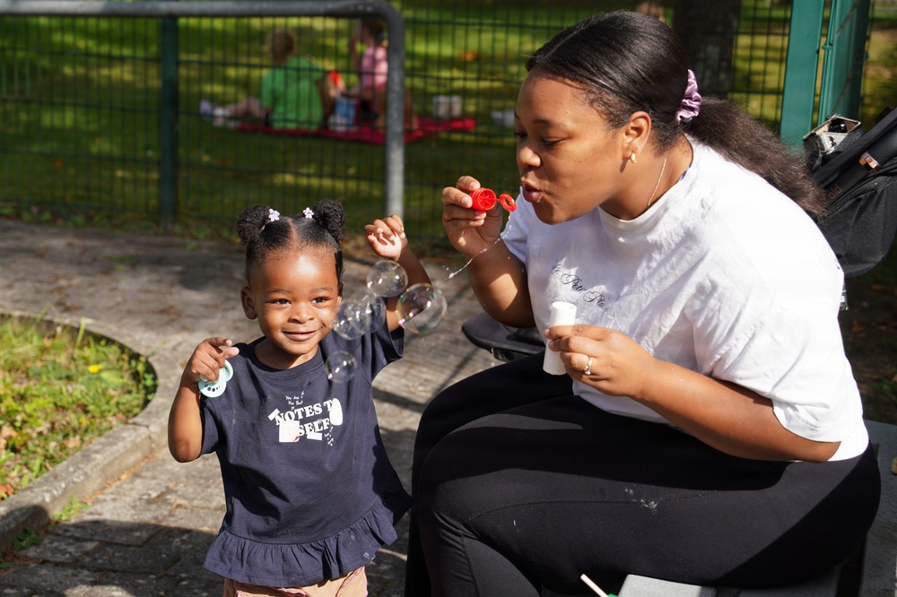 A young woman and a toddler girl are enjoying a bubble-blowing activity together at an outdoor park or playground. The woman is seated and blowing bubbles using a red bubble wand while the little girl, dressed in a navy 'Notes to Self' shirt, smiles and reaches toward the bubbles. Several bubbles are visibly floating in the air between them.