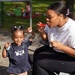 A young woman and a toddler girl are enjoying a bubble-blowing activity together at an outdoor park or playground. The woman is seated and blowing bubbles using a red bubble wand while the little girl, dressed in a navy 'Notes to Self' shirt, smiles and reaches toward the bubbles. Several bubbles are visibly floating in the air between them.