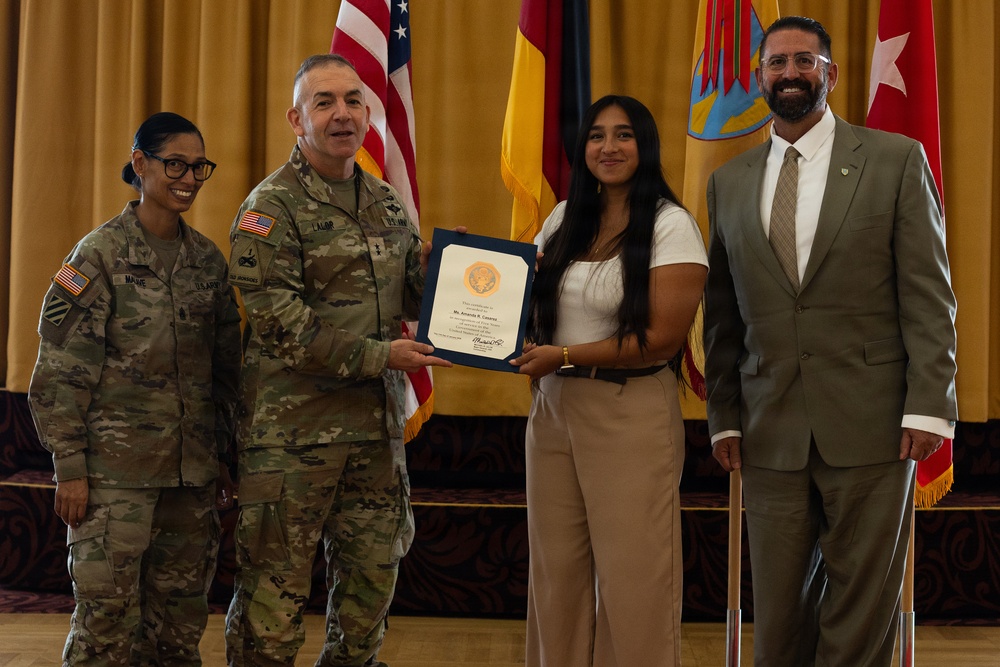 A formal military award ceremony is taking place in what appears to be a ceremonial hall. Two U.S. Army soldiers in camouflage uniforms are presenting a certificate or award to a young civilian woman, while a man in a suit stands to the right. Multiple flags, including the U.S. flag and military unit flags, are displayed in the background.