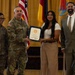 A formal military award ceremony is taking place in what appears to be a ceremonial hall. Two U.S. Army soldiers in camouflage uniforms are presenting a certificate or award to a young civilian woman, while a man in a suit stands to the right. Multiple flags, including the U.S. flag and military unit flags, are displayed in the background.