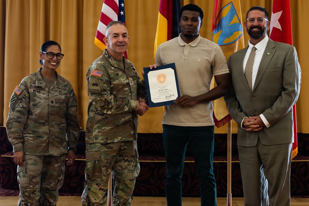 A formal military ceremony is taking place in which a uniformed Army officer is presenting a certificate or award to a civilian young man while shaking his hand. Four individuals are present: two in U.S. Army combat uniforms, one civilian recipient, and one civilian in a suit. The setting includes American and military unit flags against a gold/yellow curtain backdrop.