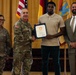 A formal military ceremony is taking place in which a uniformed Army officer is presenting a certificate or award to a civilian young man while shaking his hand. Four individuals are present: two in U.S. Army combat uniforms, one civilian recipient, and one civilian in a suit. The setting includes American and military unit flags against a gold/yellow curtain backdrop.