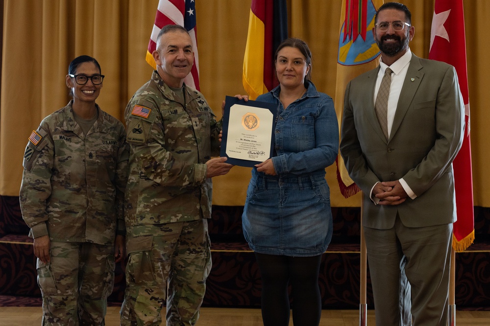 A formal military award or recognition ceremony is taking place indoors, likely on a U.S. Army installation. A uniformed Army officer is presenting a certificate or commendation to a civilian woman in a denim dress, flanked by another soldier and a civilian man in a suit. Multiple flags, including the U.S. flag and what appears to be a German flag, are visible in the background.