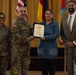 A formal military award or recognition ceremony is taking place indoors, likely on a U.S. Army installation. A uniformed Army officer is presenting a certificate or commendation to a civilian woman in a denim dress, flanked by another soldier and a civilian man in a suit. Multiple flags, including the U.S. flag and what appears to be a German flag, are visible in the background.