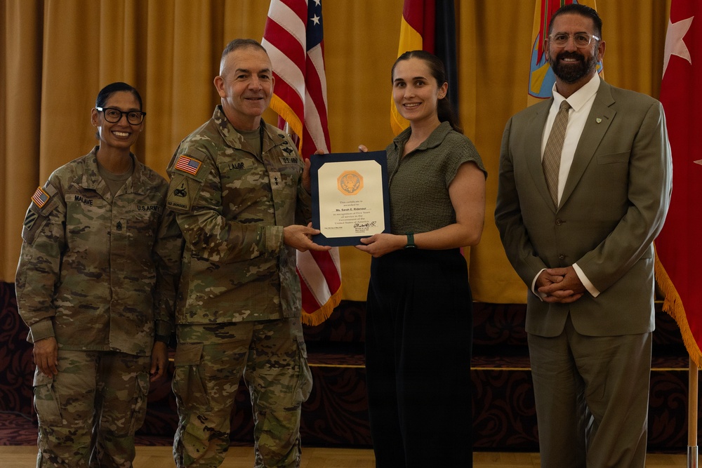 A formal military award ceremony is taking place in what appears to be a ceremonial hall. A U.S. Army officer with the name tape 'LALOR' is presenting a certificate or commendation to a civilian woman, while two other individuals — a female soldier with name tape 'MALAVE' and a civilian man in a suit — stand alongside them. Multiple flags, including the U.S. flag and military unit flags, are visible in the background.