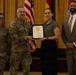A formal military award ceremony is taking place in what appears to be a ceremonial hall. A U.S. Army officer with the name tape 'LALOR' is presenting a certificate or commendation to a civilian woman, while two other individuals — a female soldier with name tape 'MALAVE' and a civilian man in a suit — stand alongside them. Multiple flags, including the U.S. flag and military unit flags, are visible in the background.