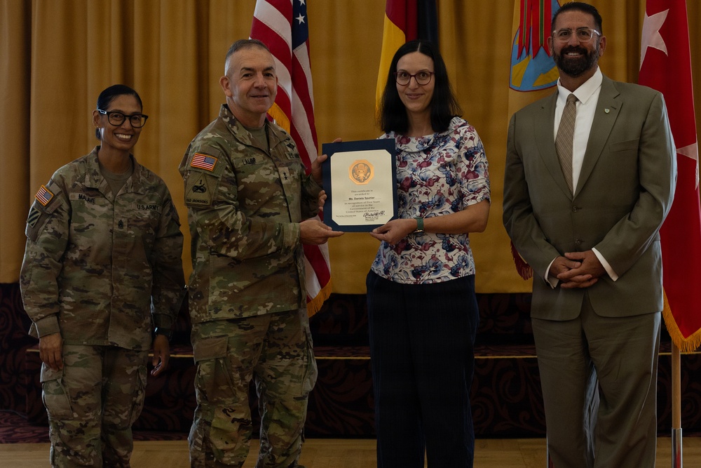 A formal military award ceremony is taking place indoors, where a U.S. Army soldier in camouflage uniform is presenting a framed certificate to a civilian woman. Three other individuals are present, including a female soldier on the left with the name tape 'MALAVE' and a civilian man in a suit on the right. Multiple flags, including the U.S. flag and what appears to be a unit or organizational flag, are displayed in the background.