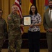 A formal military award ceremony is taking place indoors, where a U.S. Army soldier in camouflage uniform is presenting a framed certificate to a civilian woman. Three other individuals are present, including a female soldier on the left with the name tape 'MALAVE' and a civilian man in a suit on the right. Multiple flags, including the U.S. flag and what appears to be a unit or organizational flag, are displayed in the background.