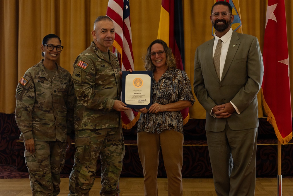 A formal military award ceremony is taking place in what appears to be a ceremonial hall. A U.S. Army soldier in combat uniform is presenting a framed certificate to a civilian woman, while two other individuals — a female soldier and a civilian man in a suit — stand alongside them. Flags including the U.S. flag and what appears to be a German flag are visible in the background.