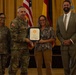 A formal military award ceremony is taking place in what appears to be a ceremonial hall. A U.S. Army soldier in combat uniform is presenting a framed certificate to a civilian woman, while two other individuals — a female soldier and a civilian man in a suit — stand alongside them. Flags including the U.S. flag and what appears to be a German flag are visible in the background.