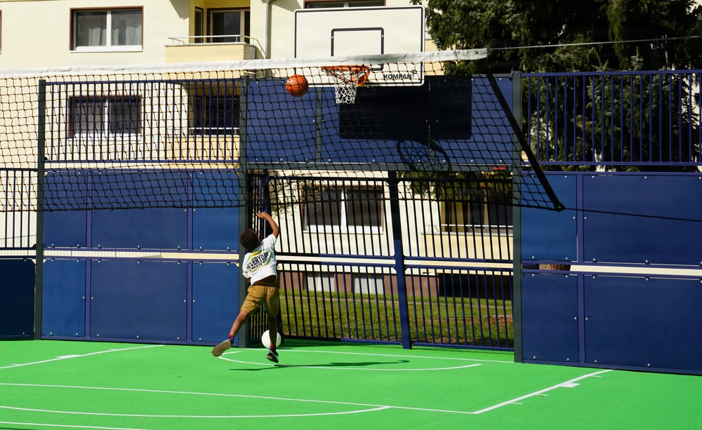 A child is playing basketball on a multi-sport outdoor court, captured mid-jump while attempting a shot toward a KOMPAN basketball hoop. The court features a green synthetic surface with white line markings and is enclosed by blue metal fencing panels with a net. A residential building is visible in the background.