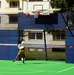 A child is playing basketball on a multi-sport outdoor court, captured mid-jump while attempting a shot toward a KOMPAN basketball hoop. The court features a green synthetic surface with white line markings and is enclosed by blue metal fencing panels with a net. A residential building is visible in the background.