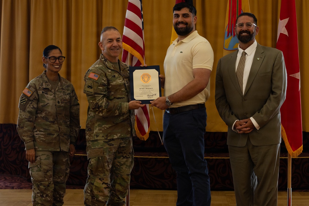 A formal military ceremony is taking place in what appears to be a banquet or event hall. A uniformed military officer is presenting a certificate or award to a civilian man in a yellow polo shirt, while two other individuals — a female soldier and a suited civilian — stand nearby smiling. American flags and a military unit flag are visible in the background.