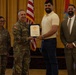 A formal military ceremony is taking place in what appears to be a banquet or event hall. A uniformed military officer is presenting a certificate or award to a civilian man in a yellow polo shirt, while two other individuals — a female soldier and a suited civilian — stand nearby smiling. American flags and a military unit flag are visible in the background.