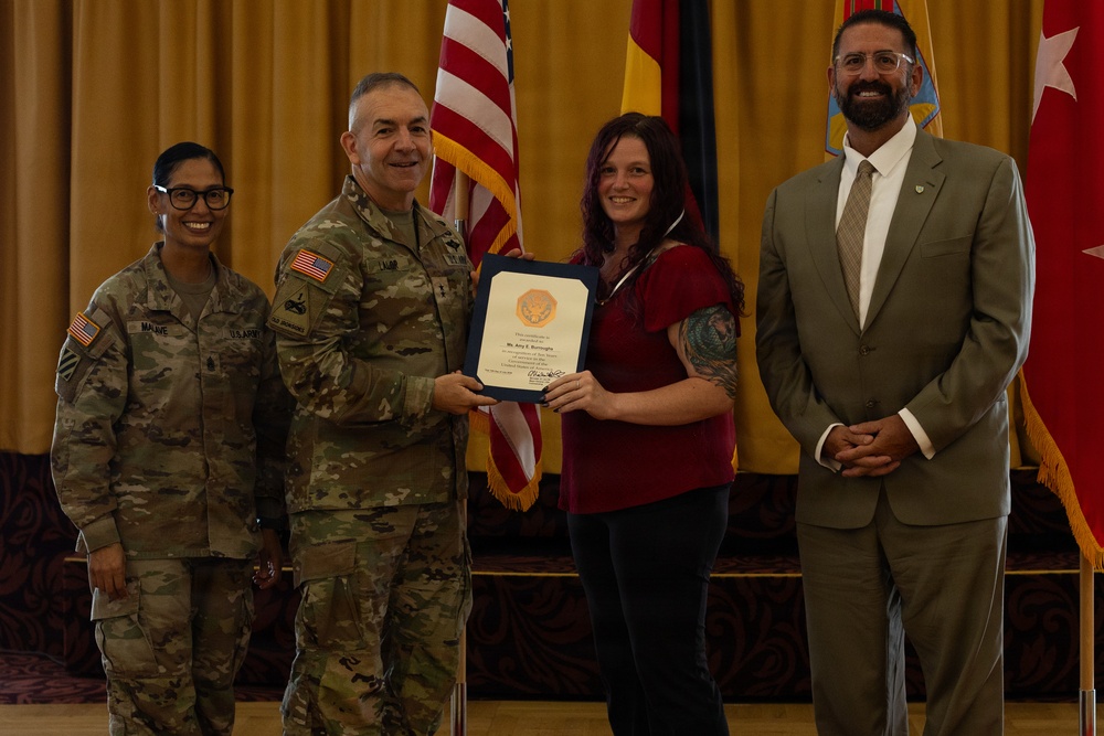 A formal military award ceremony is taking place in what appears to be a banquet or conference hall. A U.S. Army officer in camouflage uniform is presenting a framed certificate to a civilian woman, while two other individuals — a female soldier and a civilian man in a suit — stand alongside them. Multiple flags, including the U.S. flag and German flag, are visible in the background.