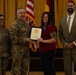 A formal military award ceremony is taking place in what appears to be a banquet or conference hall. A U.S. Army officer in camouflage uniform is presenting a framed certificate to a civilian woman, while two other individuals — a female soldier and a civilian man in a suit — stand alongside them. Multiple flags, including the U.S. flag and German flag, are visible in the background.
