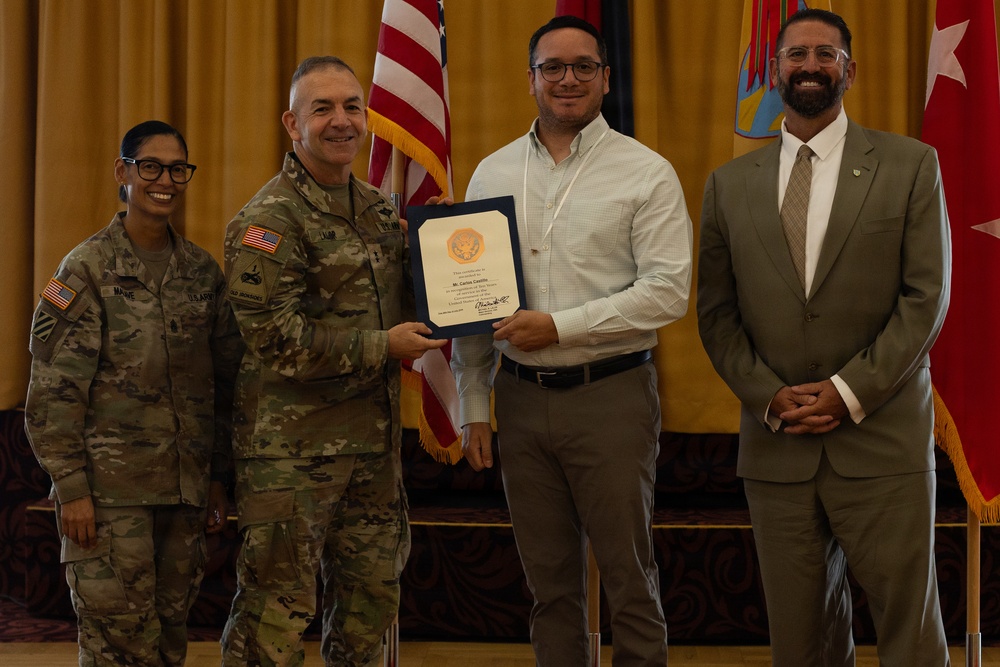A military award ceremony is taking place in a formal indoor setting. A U.S. Army officer in camouflage uniform is presenting a framed certificate to a civilian male, while two other individuals — a female soldier and a suited civilian — stand nearby. Flags including the U.S. flag and a military unit flag are visible in the background.