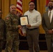 A military award ceremony is taking place in a formal indoor setting. A U.S. Army officer in camouflage uniform is presenting a framed certificate to a civilian male, while two other individuals — a female soldier and a suited civilian — stand nearby. Flags including the U.S. flag and a military unit flag are visible in the background.