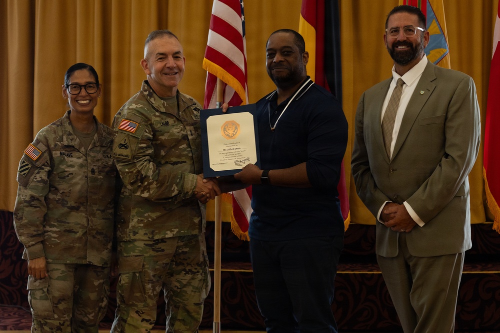 A military award ceremony is taking place in a formal indoor setting. A uniformed Army officer is shaking hands with a civilian man while presenting a framed certificate, with two other individuals standing alongside them. The setting includes multiple flags including the U.S. flag and military unit flags in the background.