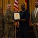 A military award ceremony is taking place in a formal indoor setting. A uniformed Army officer is shaking hands with a civilian man while presenting a framed certificate, with two other individuals standing alongside them. The setting includes multiple flags including the U.S. flag and military unit flags in the background.