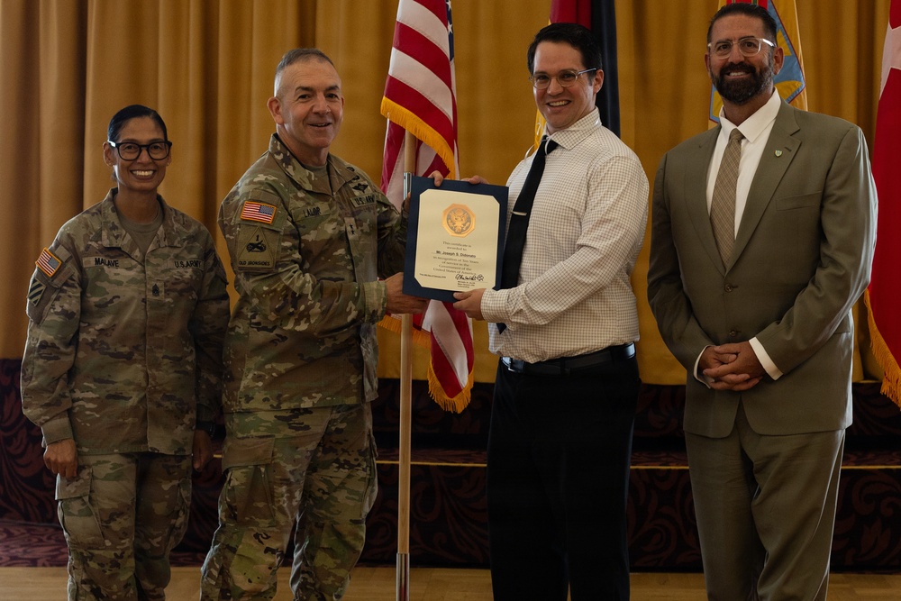 A formal military award or recognition ceremony is taking place indoors, likely at a U.S. Army installation. A uniformed Army officer is presenting a framed certificate or commendation to a civilian male, while two other individuals — a female soldier and a civilian male — stand alongside them. An American flag and additional military unit flags are visible in the background.
