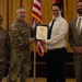 A formal military award or recognition ceremony is taking place indoors, likely at a U.S. Army installation. A uniformed Army officer is presenting a framed certificate or commendation to a civilian male, while two other individuals — a female soldier and a civilian male — stand alongside them. An American flag and additional military unit flags are visible in the background.