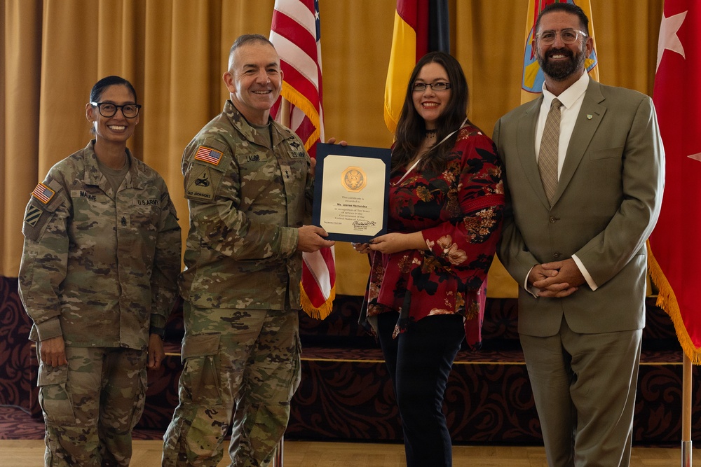 A formal military award ceremony is taking place in what appears to be a banquet or conference hall. A U.S. Army officer in camouflage uniform is presenting a framed certificate to a civilian woman in a red floral outfit, while two others stand alongside them. Flags including the U.S. flag and what appears to be a New Mexico state flag are visible in the background.