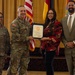 A formal military award ceremony is taking place in what appears to be a banquet or conference hall. A U.S. Army officer in camouflage uniform is presenting a framed certificate to a civilian woman in a red floral outfit, while two others stand alongside them. Flags including the U.S. flag and what appears to be a New Mexico state flag are visible in the background.