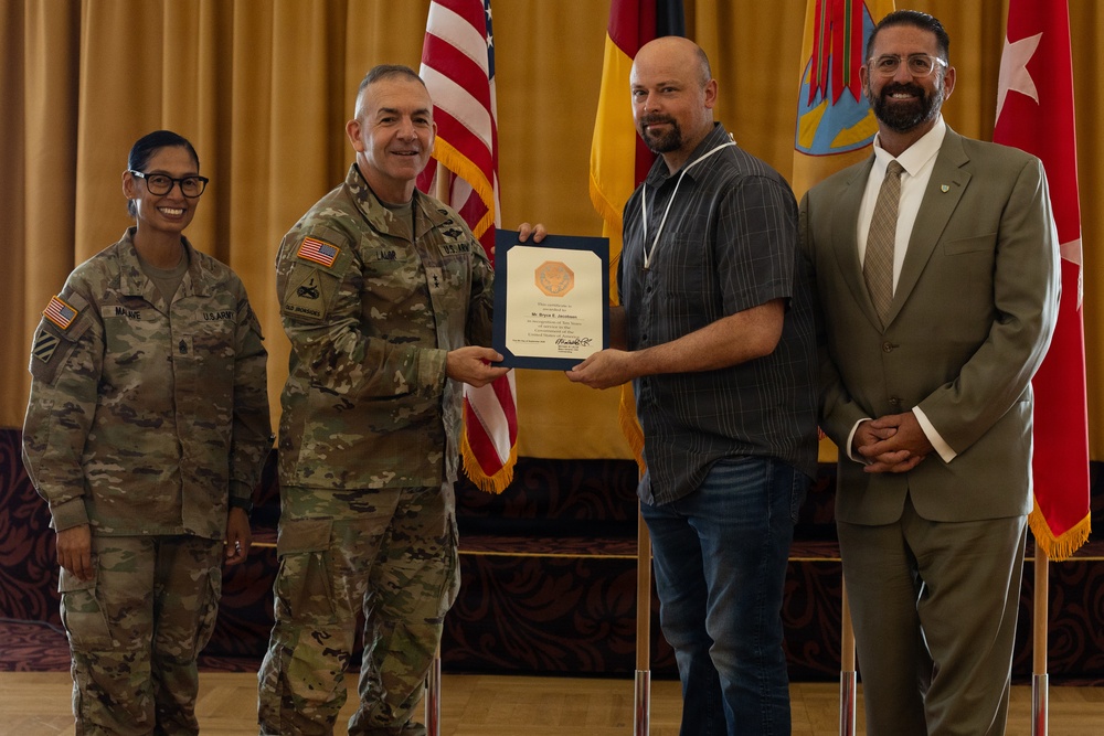 A military ceremony is taking place in which a U.S. Army officer is presenting a certificate or award to a civilian male. Four individuals are present: two in U.S. Army combat uniforms, one civilian recipient, and one civilian in a suit. The setting appears to be a formal event hall with American and military flags displayed in the background.