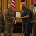 A military ceremony is taking place in which a U.S. Army officer is presenting a certificate or award to a civilian male. Four individuals are present: two in U.S. Army combat uniforms, one civilian recipient, and one civilian in a suit. The setting appears to be a formal event hall with American and military flags displayed in the background.