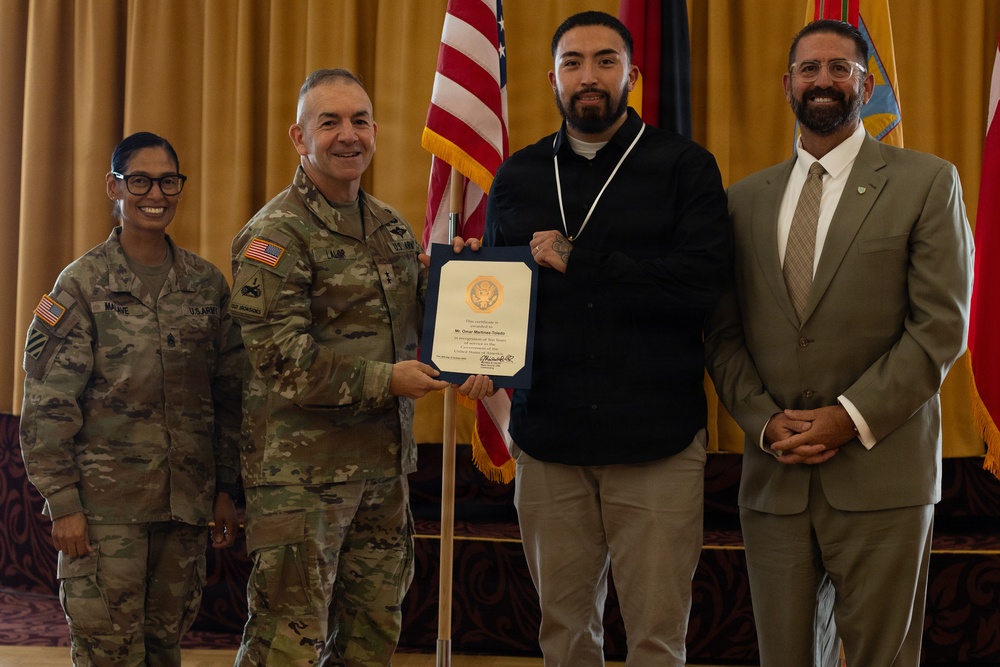 A formal military ceremony in which a U.S. Army general presents a certificate to a civilian recipient. Four individuals are posed together in front of an American flag and military unit flags in what appears to be a banquet or event hall. The civilian recipient is wearing a medal around his neck and holding the blue-framed certificate.