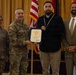 A formal military ceremony in which a U.S. Army general presents a certificate to a civilian recipient. Four individuals are posed together in front of an American flag and military unit flags in what appears to be a banquet or event hall. The civilian recipient is wearing a medal around his neck and holding the blue-framed certificate.